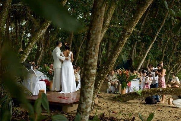 A wedding ceremony on the lodge's beach — bride and groom on a petal-strewn wooden platform under the beach almonds, guests seated at the treeline