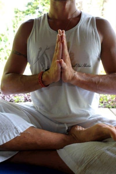 Hands at heart centre — seated practice with the garden green behind the shala