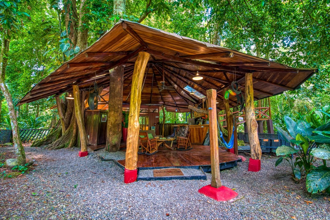 Open-air rancho at ground level — red wooden columns, thatched roof, hammock and bench